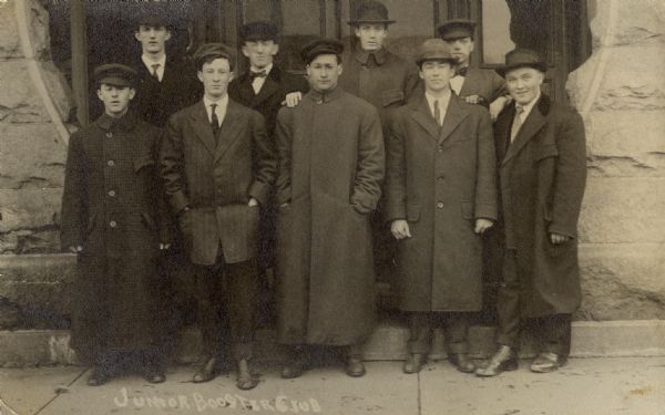 Group portrait of nine young men, in coats and hats, standing together on a sidewalk in front of the entrance to a building. Caption reads: "Junior Booster Club."