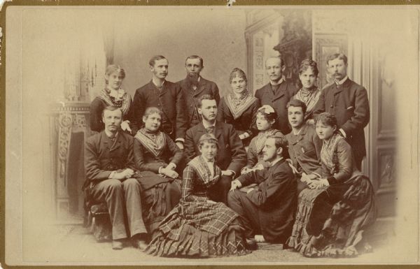 Group portrait in front of a painted backdrop of fifteen well-dressed men and women. Caption identifies them as members of the Pecan Club, attending the Sun Prairie Snowbound Party. The people are identified as: Emma Wetherbee, Ella Harris, Kate Paul, Mame Bunn, Carrie Kellogg, Grace Clark, Frank Clark, Albert Dexter, Chas. Miller, Ed Paul, Brigham Bliss, Henry Schuette, Will Lyon, Chas. Kellogg, and "Riley."