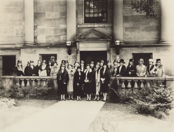 Group portrait of twenty-six women in hats and coats posing outdoors in front of a building with a stone balustrade and columns framing the entrance. The woman in the center of the back row is believed to be Laura Chapman Miller.