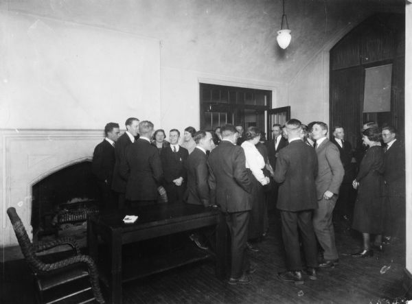 Group of men and women visiting together in a large room. A table and chair are on the left near a fireplace.