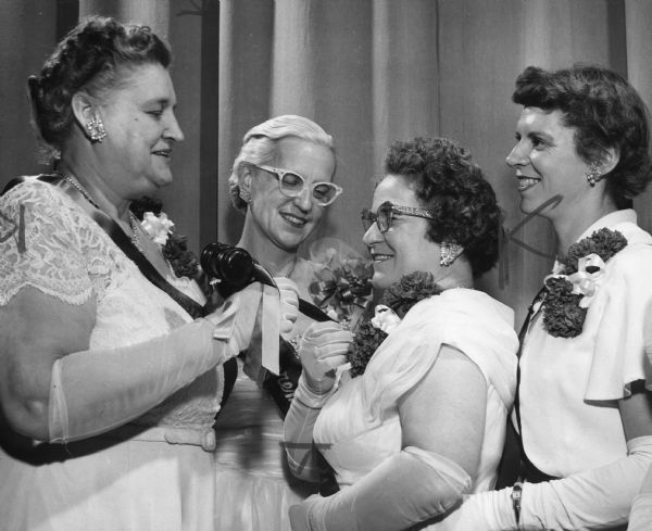 Four women wearing gowns and are gloves. One of the women is handing a gavel to another. Caption reads: "New officers of Emblem Club No. 44 of the Milwaukee Elks were installed at a meeting Thursday night of the Elks Club. From left are Mrs. William D. Westerman, 2479 N. 66th St., Wauwatosa, past junior president of the club; Mrs. Evelyn Hummel, Janesville, past supreme president and installing officer, and Mrs. Eugene Leister, 5268 N. 38th St., new president."