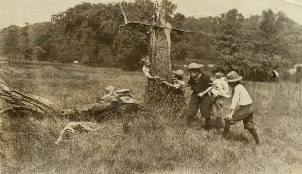 Seven children, some wearing hats and holding toy guns, playing outdoors at a gunfight. One child is hiding behind a tree stump, another child is hiding behind a felled branch, and one child is playing dead in the field.