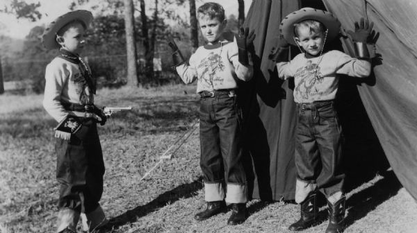 Three boys are dressed in cowboy hats, gloves, boots, and t-shirts featuring Roy Rogers and his horse Trigger. One boy is holding a toy pistol on the other two, who have their hands up while standing outdoors in front of a tent. A caption identifies the boys as Bruce, Bill, and Bob Bryan.