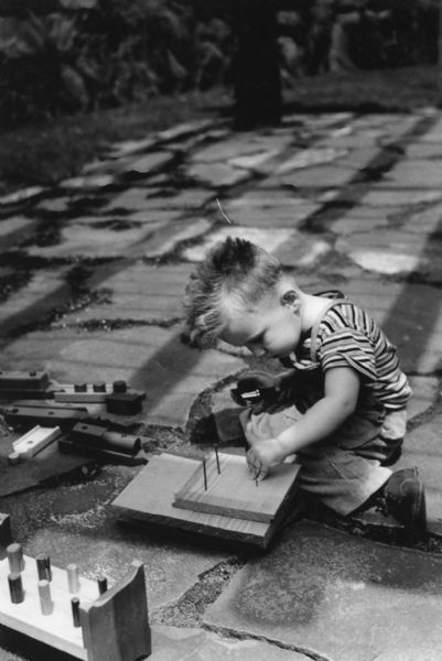A boy is sitting on the ground and getting ready to hammer nails through two boards.