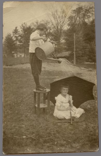 A boy is standing on a small table and pouring water, out of a watering can, onto a girl sitting in the grass holding up an umbrella.