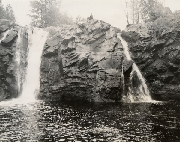 View from below towards two waterfalls along a rock formation. A person is sitting on the rock outcropping between the falls.