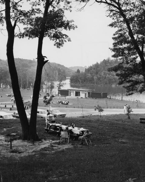 Elevated view of a park scene near Cox Hollow Lake, at Governor Dodge State Park. In the foreground, a man is grilling at a picnic table, while two women are sitting nearby. Cars are parked in a parking lot. In the distance, several people are sitting or playing on the lawn, and a shelter or bathroom facility is near the lakeshore.