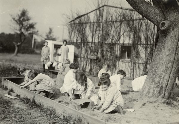 Several children playing in a sandbox, which has a tree growing from its center. In the background and out of focus, two women are standing near what could be an open shed. A building is behind them is covered in vines.