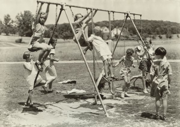 Several girls are playing on a swing-set, which has board seats and rings at various heights. A caption identifies the photograph as having been taken in Shorewood Hills.