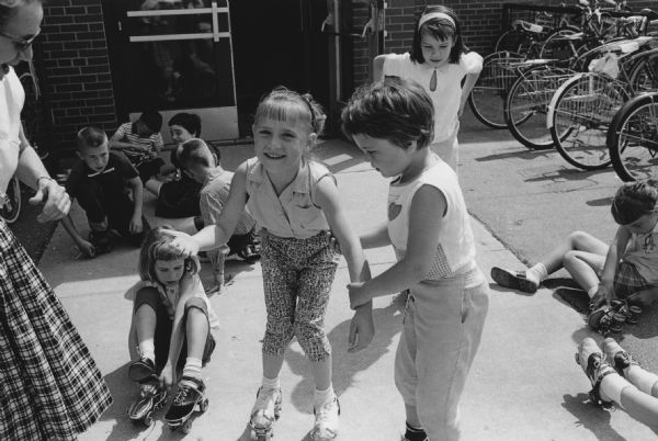 Several children are putting on roller skates outside a school door. Bicycles are parked in the background and a woman is partially visible in the foreground. A caption identifies this as Midvale School Kindergarteners, Madison