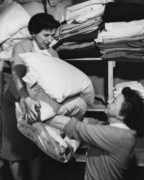 One woman is handing folded linens to another woman while they are standing in a large closet. Caption reads: "The linen closet received a thorough going over by Mrs. Jerome H. Schwarz (left), 8316 N. Santa Monica Blvd., Fox Point. She is among about 60 members of the outing association which helps support the camp."