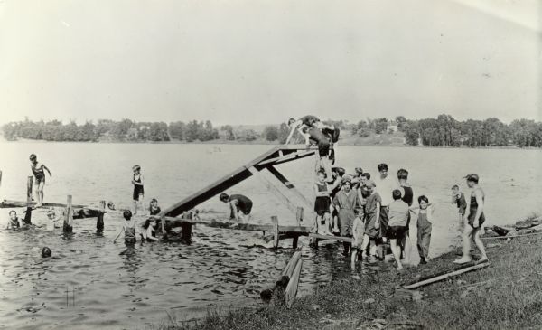 View from shoreline of a lake towards several children climbing or standing on a wooden pier. On the side of the pier is a wooden slide that angles into the water. Some of the children are swimming in the water, and children are climbing up the ladder to the top of the slide.