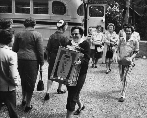 Women are walking to and from a parked bus, some of them carrying boxes and bags of books. Caption reads: "Mrs. Theodore L. [Ann] Albert, 404 E. Apple Tree Rd., Fox Point, carted a load of books from the bus on which the women made the short trip to Delafield. About 35 workers participated in the annual clean up operation. The camp will be opened on June 22."