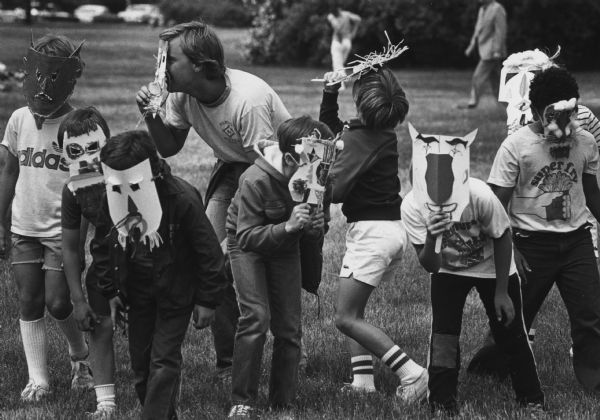 A man and several children, all wearing handmade masks, playing in a field. Caption reads: "Jay Wigdale, who runs a private day camp, brought his charges to the Lakefront Festival of Arts. The children, and Wigdale, romped with the masks they made Friday at the Mask Center, part of the Family Center at the festival."