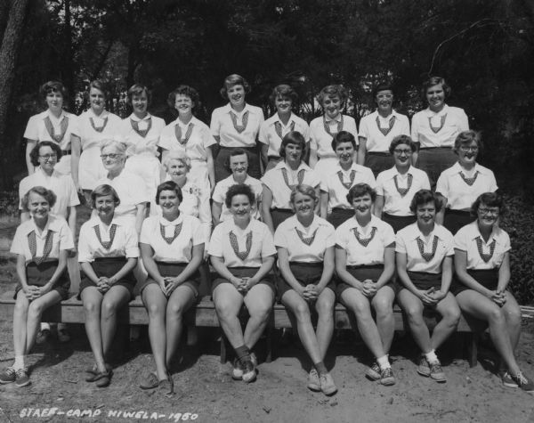 Three rows of women are posing outdoors for a group portrait. They are wearing uniforms for a Camp Hiwela.