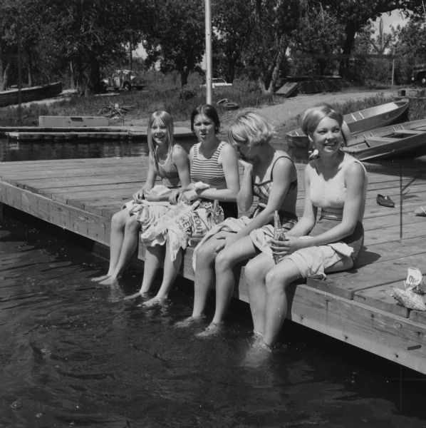 Group portrait of four young women sitting on the edge of a dock with their feet in the water. One woman is holding a Pepsi bottle. In the background, boats are pulled up on the shoreline. Caption reads: "Beach, dock, and spring-fed fish pond make Bay City a wonderful place for young people to spend their summers. Here are Jill Johnson, Kathy Dougherty, Nanci Evans and Lynn Johnson."