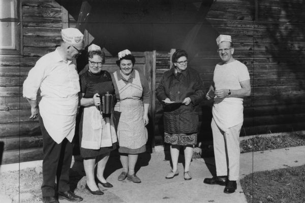 Group portrait of two men and three women standing outside a log cabin. One woman is holding a coffee carafe, another is holding some papers, and one man is holding a spatula. The men are wearing paper hats for Oak Park Dairy Foods. Caption reads: "Directors of the Eau Claire County Youth Camp assist at a recent pancake supper which drew about 700 people. From left are: Henry Kolka, Olga Martin, Dorothy Kopplin, Sarah Weippert and George Jevne."