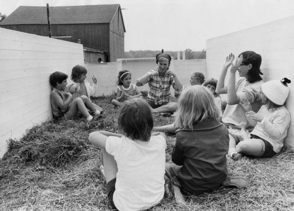 Two women and several children are sitting in a low-walled enclosure filled with hay. In the background is a barn.