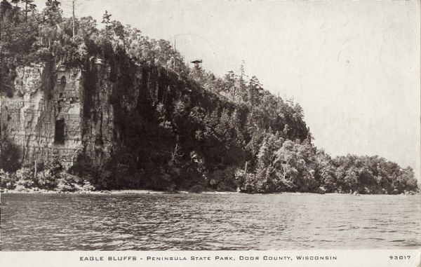 Photographic postcard view across water towards Eagle Bluffs along the Lake Michigan shoreline. This postcard was mailed to Miss Harriet Shepherd, 1144 Race St., Denver, Colorado. On the reverse is written: "Going home tomorrow. We exhibited at the Hobby Show here & sold a lot of things at the best gift shop here. Will write later. L.S." 