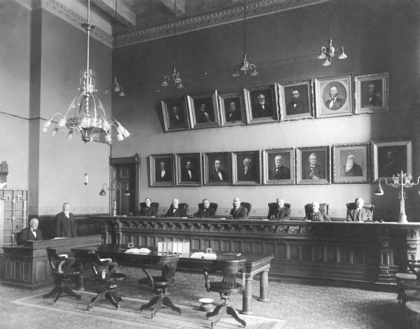 The Wisconsin State Capitol courtroom. Judges are sitting in seats below a wall of framed portraits.