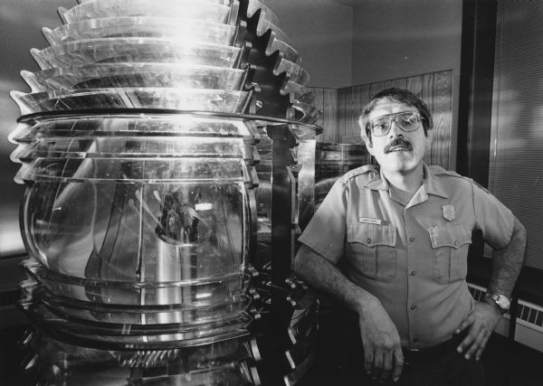 Portrait of a uniformed man leaning on a large lighthouse lens. Caption reads: "US National Park Ranger Jim Mack (above), leaning on a giant fresnel lens that was once a lighthouse beacon, said volunteer lighthouse keepers are 'a very enthusiastic bunch.'"