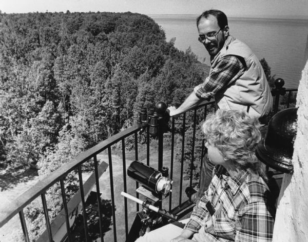 View of a man and a woman looking over a railing, with binoculars and a telescopic lens at hand. Caption reads: "Light keeper John Foote and researcher Lauri Scott kept track of eagles' nests from the Outer Island lighthouse."