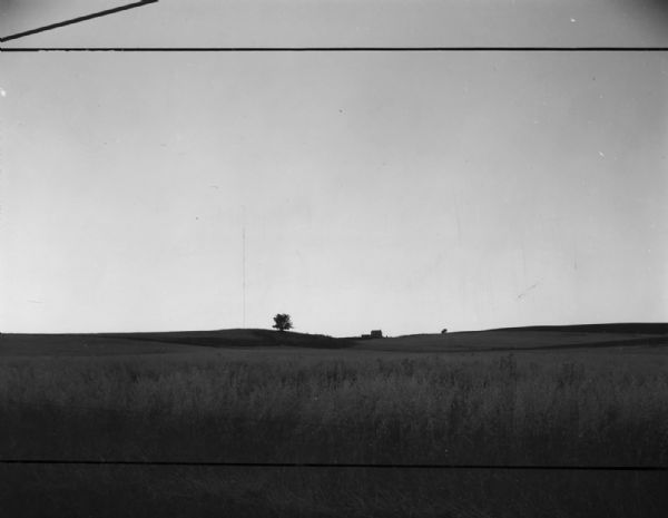 View across a field, with a small hill in the distance. One tree is on the hill, and a building is nearby. Behind them, a tower extends into the sky. Caption reads: "Madison (vicinity) July 30 '60. Television station tower at southwest edge of the city."