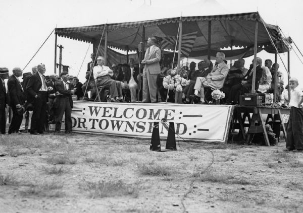 Several men are sitting on a raised platform with an awning overhead, and a banner along the front that reads: "Welcome Dr. Townsend." One man is speaking into a microphone and several men standing on the left are listening. On stage, Dr. Francis Townsend the fourth man to the right of the speaker. Dr. Townsend originated the "Townsend Plan," which would guarantee a monthly pension to all retired persons age 60 or older. His plan was a precursor to the Social Security system.