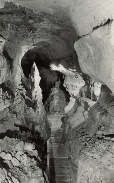 View of a large interior cave room. There is a wooden bridge and a lighting fixture. Caption reads: "Hall of Travertine, a single room 350 feet long with the ceiling 30 feet from the floor was excavated by a solution action of water acting through the ages on solid limestone walls. In the distance, left center, towers Statuary Ridge, a massive cave onyx deposit built up on the floor of the cave by slow dripping water. Cave of the Mounds, Blue Mounds, Wis, U.S. Highways 18 & 151 twenty five miles west of Madison, Wis."