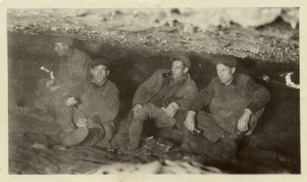 Group portrait of four men sitting inside a cave with a low ceiling. One man is holding a flashlight, and one man is smoking a pipe. A caption identifies the location as Castle Rock Cave, Town of Castle Rock, Grant County, Wis.