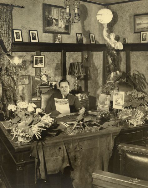 View of a man sitting behind a desk in a richly decorated office. He is holding a pipe and a Froedtert Service market report. Kurtis Froedtert was head of the Froedtert Grain and Malting Company, at its time the largest malting company in the world.