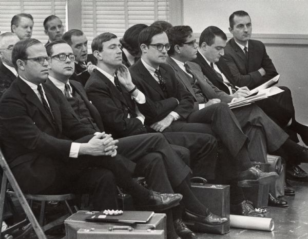 A group of men in suits are sitting on folding chairs, looking off-camera. One man is taking notes. Some of the men have briefcases next to their chairs. Caption reads: "Lined up in the front row, officials and representatives of Laird, Inc., viewed the proceedings at Tuesday's annual meeting of stockholders of Jacobsen Manufacturing Co. Third from the left is Oliver A. Kimberly, Jr., senior vice-president of the New York investment banking firm which sought Jacobsen control.