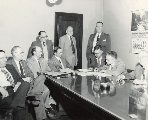 Several men are standing and sitting around a desk, where one man is signing a document. A calendar for the Norris Wholesale Corporation is on the wall behind him. Another man is holding a cigar and looking at the camera. Caption reads: "Reedsburg, Wis. April 1954. Signing agreement making Reedsburg Mill an independent mill. Mill was previously Appleton owned."