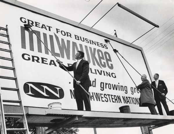 Three men are putting up a billboard sign. Two of the men are wearing suits and the third is wearing an overcoat. Caption reads: "A billboard promoting Milwaukee as "Great for Business Milwaukee Great for Living and Growing Greater," was posted at 5205 S. Howell Av." At "Work" (left to right): Ald. George Whittow, chairman of the industrial development committee; T. Parker Lowe, president of the Northwestern National Insurance Co., which is erecting five of the signs, and T.L. Mulchay, vice-president. More than 250,000 stickers carrying the slogan are being distributed to promote industry.