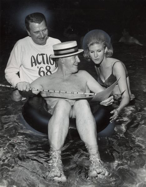 A man sitting in an inner tube and wearing swim trunks and a boater hat is using a cane to point to a document, which he is showing to a woman standing in the water next to him wearing a bikini. A man is sanding behind him wearing a sweatshirt that reads "Action '68." Caption reads: "'SOFT' WATER MEETING — Bruner Corp, a manufacturer of water conditioning equipment, held a poolside sales get-together Monday during its annual sales meeting at the Pfister hotel. Checking the 'figures' with Jan Jorden of the company promotion staff, were, from left, James L. Bruner, vice-president, and Ted W. Bruner, president."