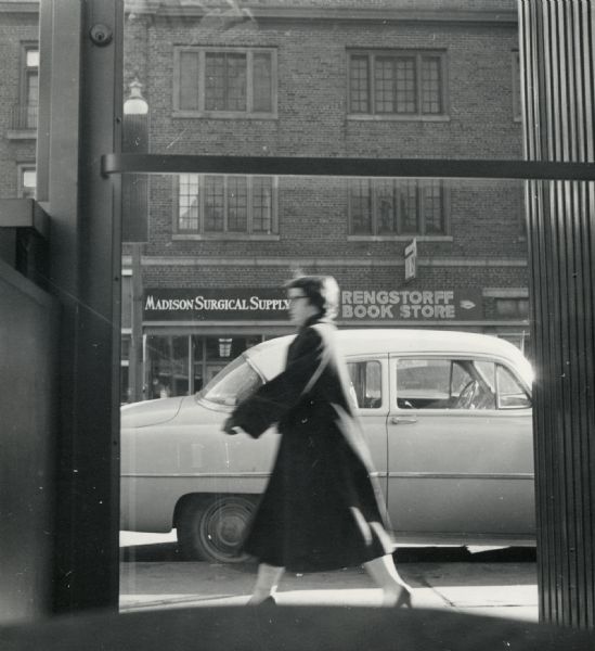 View through a glass door towards a woman walking on the sidewalk past a parked car. On the opposite side of the street are signs for Madison Surgical Supply and the Rengstorff Book Store.