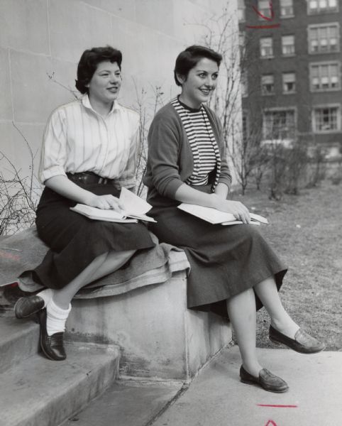 Two women are posing on the steps outside a building. Both have open books on their laps. Caption reads: "The month of March began on a good note Thursday in Milwaukee. Mary Hickey (left), Willman, Minn., and Mary Kilgallen, Chicago, Marquette university students, enjoyed the mild weather on the sunny side of the Marquette library."