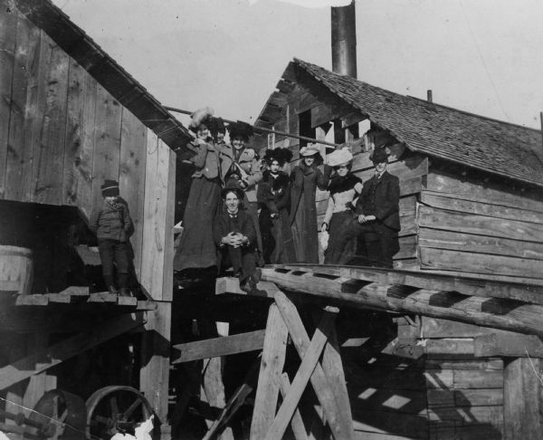 Five women, three men, and a boy are posing on a wooden bridge between two wooden structures. Caption reads: "Edgar, Wis. Yard of Tom Hill's brick factory."
