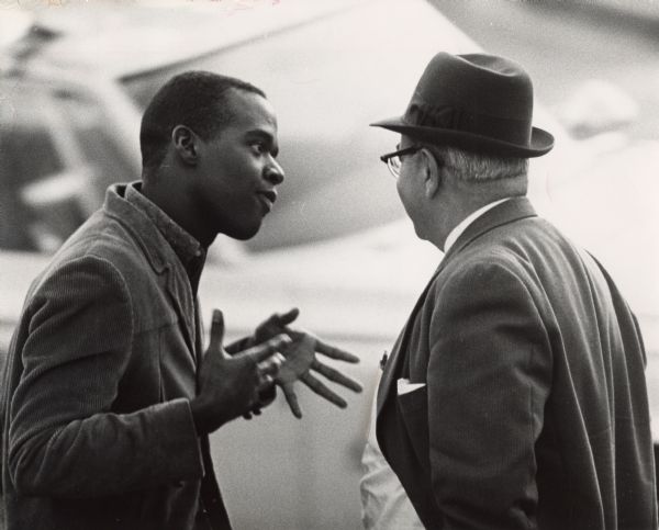 A young man on the left is talking and gesturing to an older man on the right who is partially turned away towards an automobile in the background. Caption reads: "Hands played an eloquent role in a discussion of religion as Lee Benefee (left), a seminarian, talked to a passer-by on the street, not far from St. George's Episcopal church."