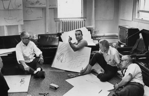 Four men are sitting or reclining on the floor in a room, surrounded by large sheets of paper and art supplies. One man is propsping up sheets of paper with drawings on it and is gesturing with his thumb to the other men. Caption reads: "In group therapy sessions such as this one in the Naval Chaplain's School, Newport, R.I., officers draw pictures and lead each other around blindfolded to learn self-awareness and group encounter techniques for better personnel management. Cmdr. Oliver Wetzel (second from right) is formerly of Mequon, Wis."