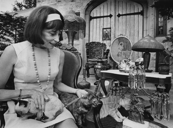 A woman is sitting in a chair with three kittens on or near her lap. A number of home decorations and pieces of furniture are nearby, and a wooden church door is in the background. Caption reads: "Three kittens in the lap of Mrs. Marshall K. Field, 2412 E. Beverly Rd., Shorewood, upstaged a collection of wares which will be sold at the Strawberry Fair to be held at St. Mark's Episcopal Church from 11 a.m. to 5 p.m. on September 12. The fair will include an art and crafts show with 30 artists exhibiting and selling their wares and a 40 section Flea Market. Neighborhood merchants will also have booths. Bratwursts, hot dogs and corn will be roasted and, of course, strawberries will be served--with ice cream. The fair, to be held in the field north of the church, is being sponsored by the women of the church. Proceeds will go to St. Mark's. And the kittens will go to the person or persons who can't resist buying them."
