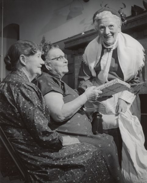 A woman in costume is smiling and stands near two women who are sitting. One of the women is holding a wrapped present. Caption reads: "Mrs. Santa Claus visited the Golden Age club party Tuesday night at the Wisconsin Avenue school. Mrs. Susana Powell (left), 81, of 542 N. 13th St., and Mrs. Louise Maegli, 82, of 2840 W. Wells St., received gifts from Mrs. Clara Peterson of 1918-A E. Webster Pl."