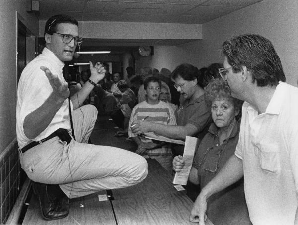 A man wearing a headset is crouching on the left on a desk or table, while a crowd of people are standing along a hallway. Caption reads: "Auctioneer Joel Cielak, of LiquiTec of Milwaukee, talked with bidders at the former nursing home [Northview Home], 25042 W. Northview Rd., Town of Pewaukee."