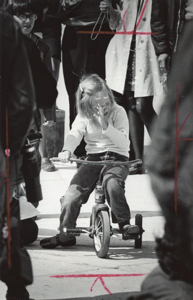 A young girl is among a crowd of people, sitting on a tricycle and crying and wiping her face. Caption reads: "ALL ALONE — Little Kim Whitham, 4, of Waukesha was having lots of fun at a campus carnival Tuesday until the cheering and shouting by college students frightened her. Kim's mother is a student at the University of Wisconsin-Waukesha. The carnival was the first for the new campus.