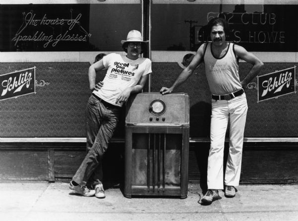 Two men are standing and leaning on a large radio outside the 602 Club, a popular Madison bar. Robin Carnes (left) is wearing a hat and a t-shirt advertising Great Big Pictures ("We blow up anything!"), a large-format printing company. M.J. Paggie (right) is wearing a sleeveless t-shirt with the 602 Club's motto: "The House of Sparkling Glasses" printed on it, in the same font as the neon window sign of the same wording. The Schlitz beer logo appears twice on the wall. Another neon sign reads "602 Club" and "Here's Howe," in reference to bar owner Dudley Howe.