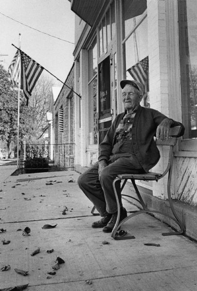 View across sidewalk towards a man sitting on a bench outside a storefront with a screen door that has a sign on it for Purity Bread. An American flag is hanging from a wall-mounted pole on the side of the building, and another flag is on a flagpole behind it. Leaves are scattered on the ground. Caption reads: "Occupying a bench on Main St., C.L. Shearer, 84, who has lived in or around the village all of his life, relaxes among the 'Americana; and the American flags of Eagle."
