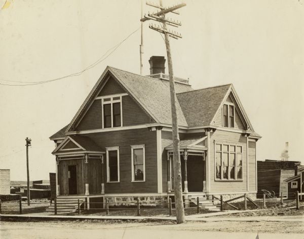 View across street towards a two-story building, with a sidewalk, low fence and a telephone or power pole in front. There is a widow's walk and a chimney on the roof of the building. Stacks of lumber are in the background on the left and right.