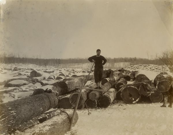 View of a man standing on a log among a large pile of logs spread out on the snow-covered ice of a lake. He is leaning on the handle of what may be a cant hook, and he has a scarf tied around his waist. Other tools are in front of him. There are a pair of boots standing in the snow on the far right, a coat laying on a log nearby, and a pair of gloves are sitting on top of a log on the far left. Low hills and trees are in the background.