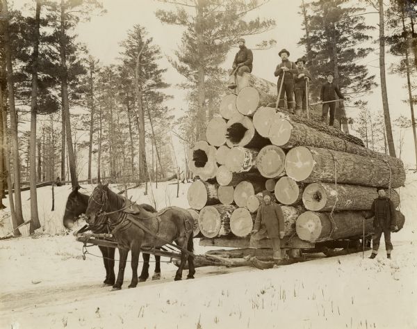 Six men are posing on and next to a large stack of logs, which are piled on a sled drawn by two horses. The men are all carrying cant hooks. The initials "WA" are carved into one log.