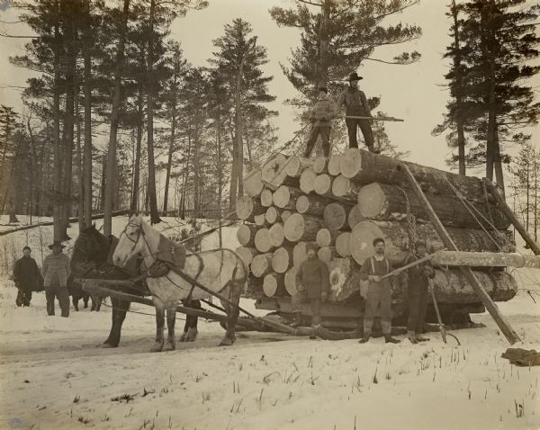 Five men are posing on or near a tall stack of logs on a sled, with more logs being added. Three of the men are carrying cant hooks, and two horses are yoked to the jammer. Two more men are standing in the background.
